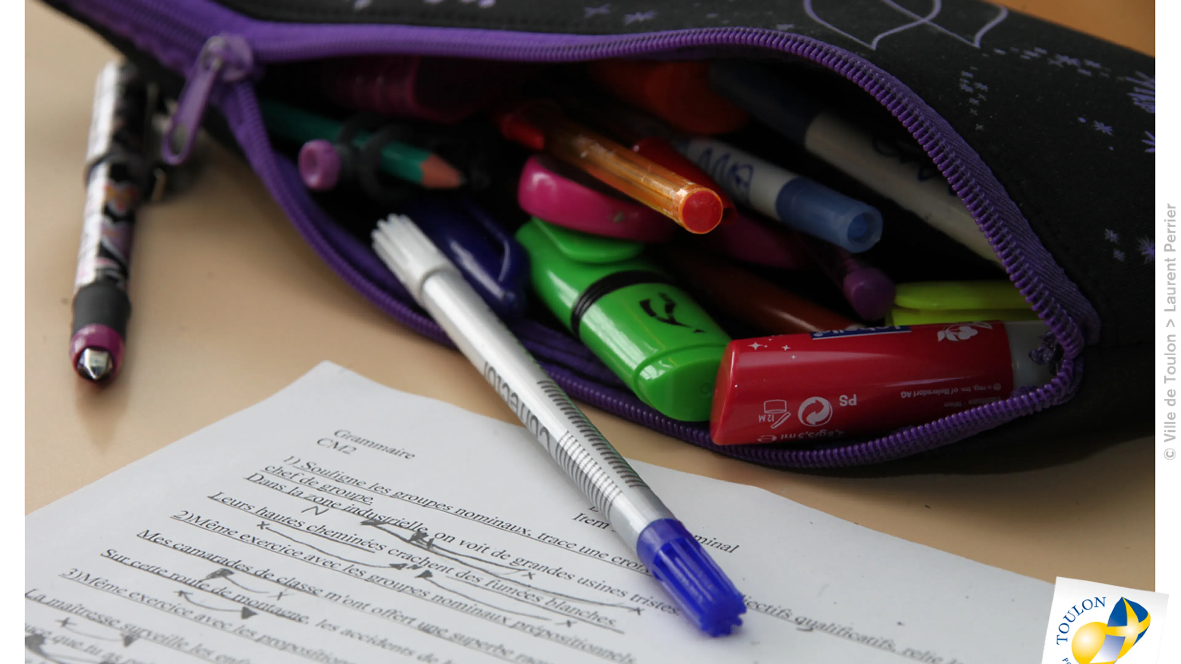 Photographie d'une trousse d'école ouverte sur un bureau, laissant tomber un stylo sur une feuille