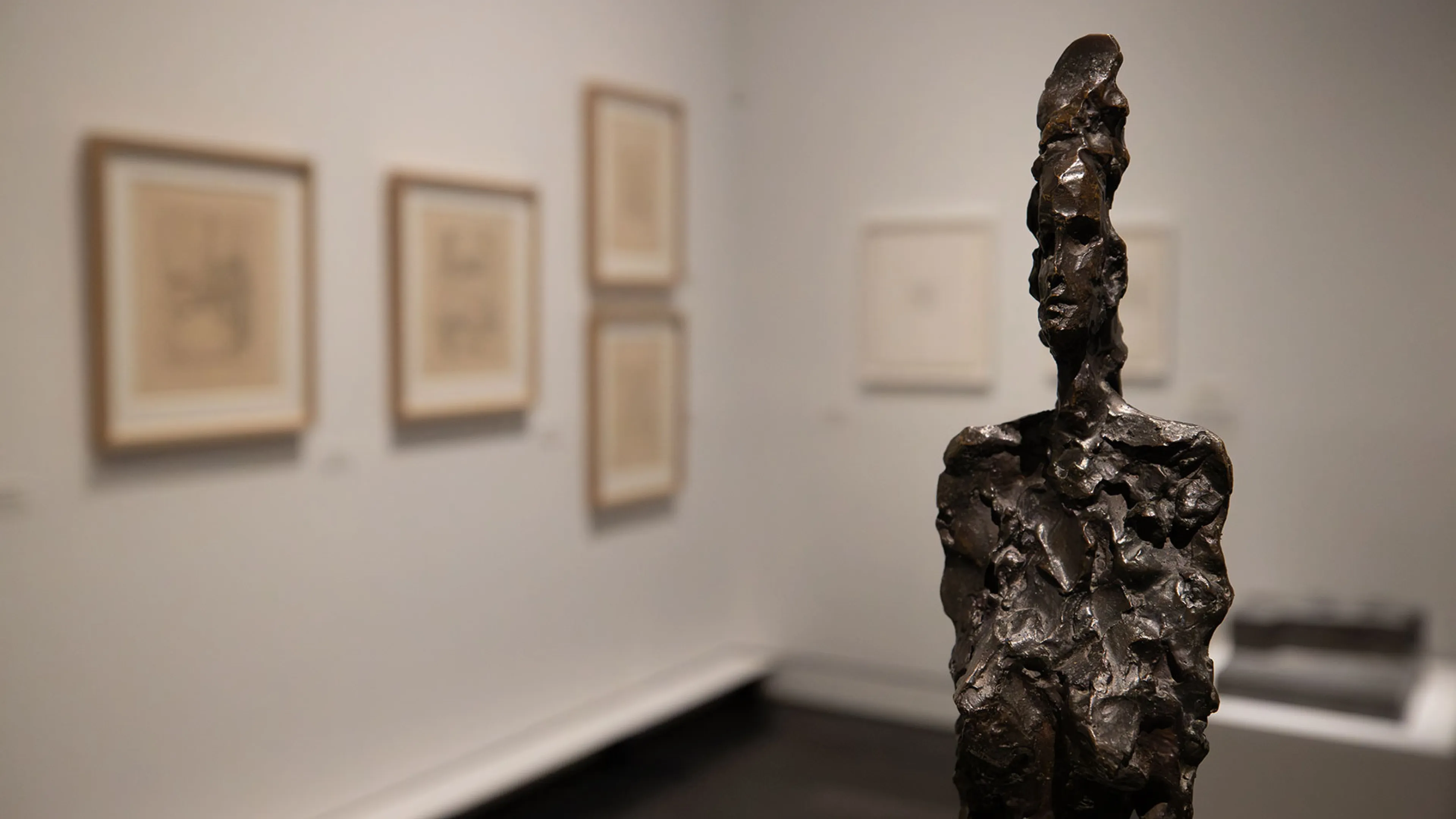 La photographie montre au premier plan la sculpture intitulée La Femme haut chignon de Giacometti et en arrière plan des gravures réalisées par l'artiste suisse.