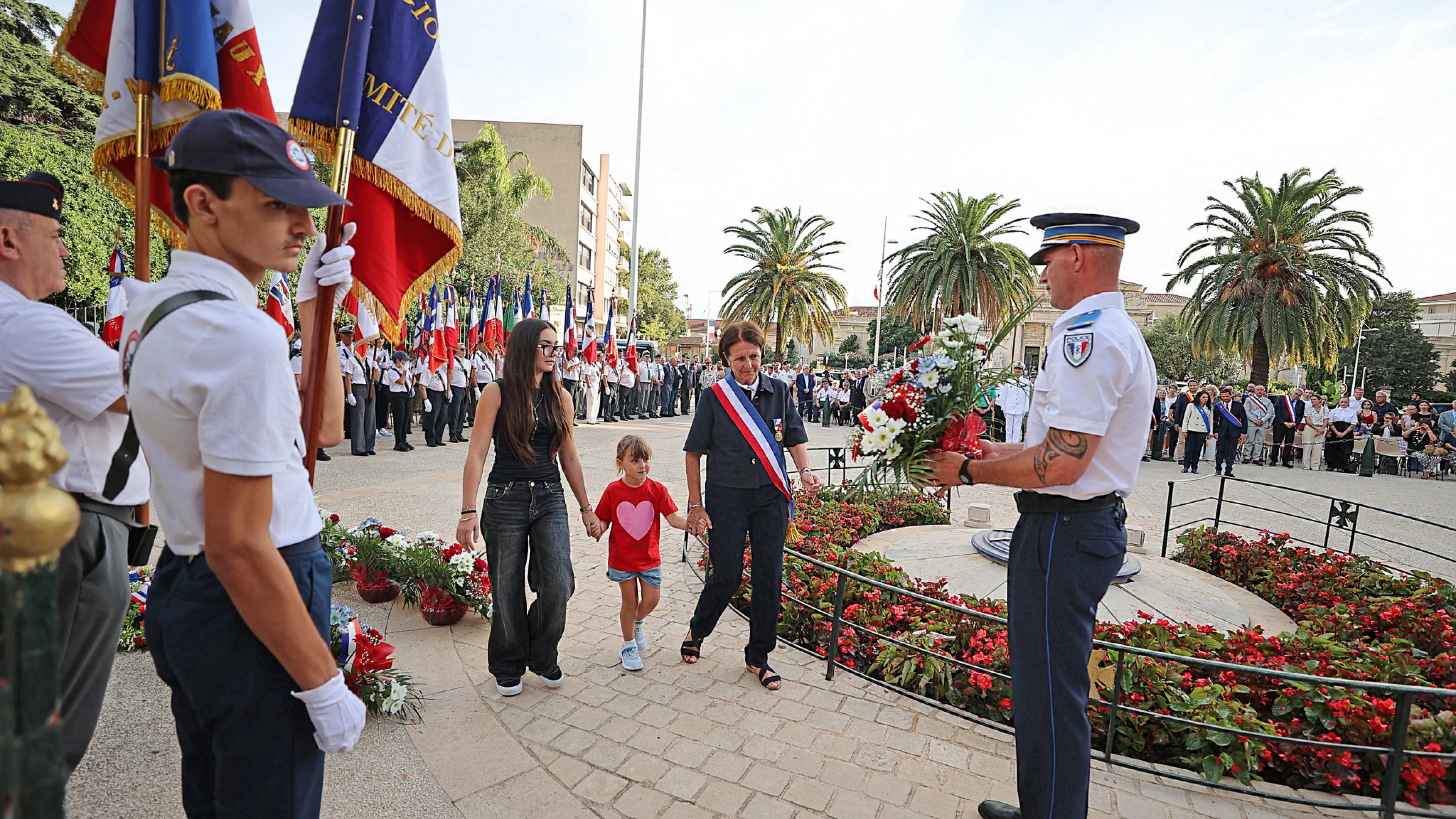La photo montre Josée Massi, maire de Toulon, entourée par deux enfants déposer une gerbe au monument aux morts de la place Gabriel Peri à l'occasion du 82e anniversaire de la libération de la ville.