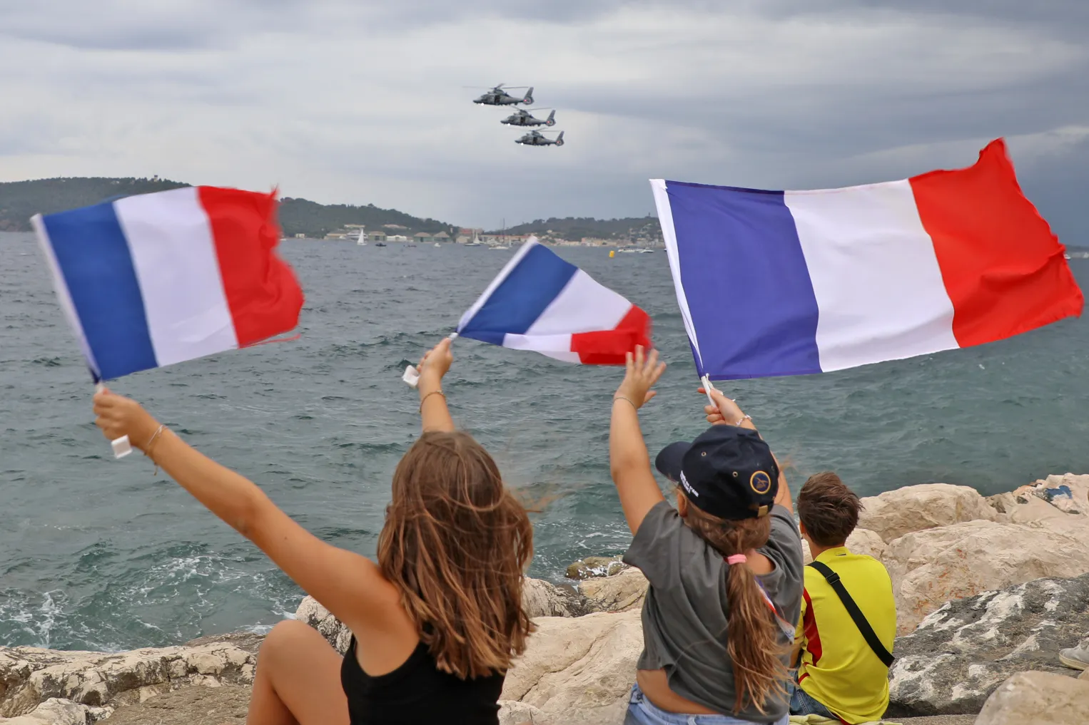 Photo : deux jeunes filles, de dos, brandissent un drapeau français, dans le ciel des avions de la Patrouille de France