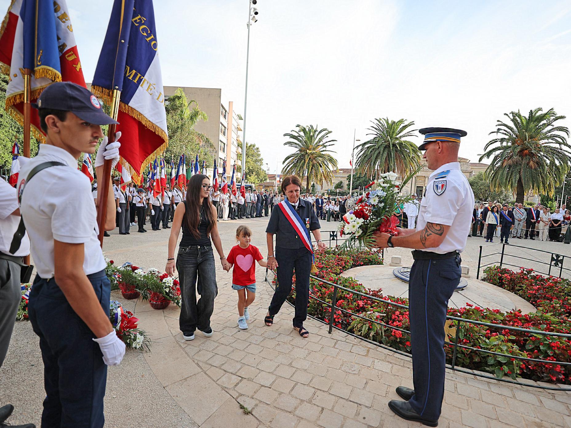 La photo montre Josée Massi, maire de Toulon, entourée par deux enfants déposer une gerbe au monument aux morts de la place Gabriel Peri à l'occasion du 82e anniversaire de la libération de la ville.