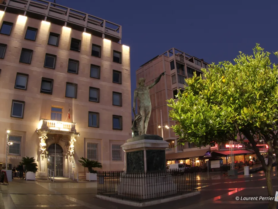 Photographie de nuit de la statue du génie de la navigation et de la Mairie d'honneur éclairée sur chacune des colonnes de sa structure, au loin derrière la statue.