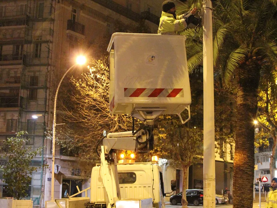 Photographie de nuit montrant un homme équipé d'un gilet jaune sur une plateforme en hauteur, tenu par un camion. L'homme est en train d'installer un système d'éclairage autour ou sur un lampadaire de la voie publique
