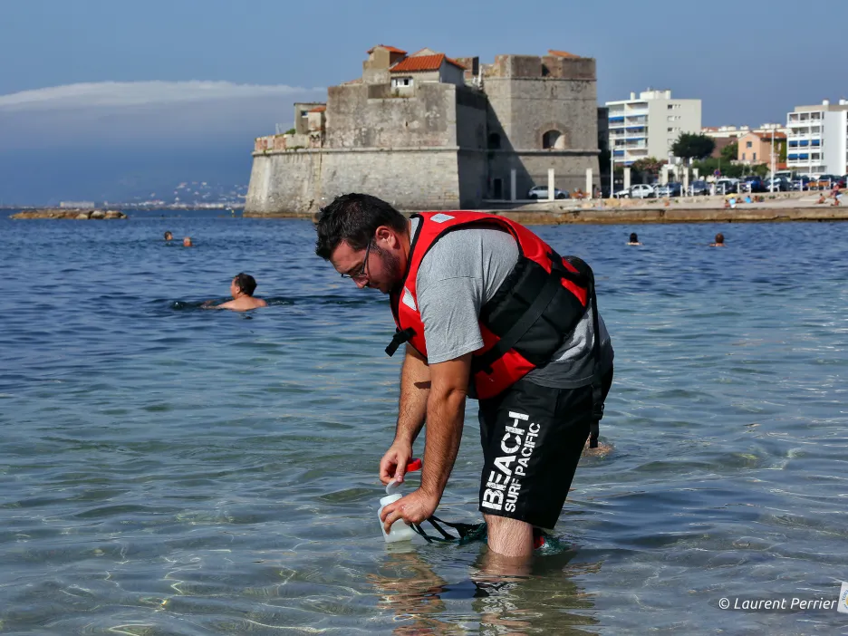 Photographie d'un agent contrôlant la qualité de l'eau des plages toulonnaises. Sur la photo, ce dernier est équipé d'un gilet rouge et on peut le voir se baisser muni de son analyseur d'eau