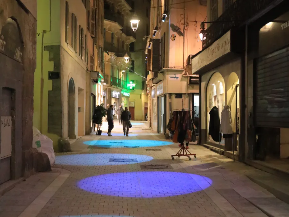 Photographie de nuit montrant une partie de la Rue des Arts : sur le sol des projections de lumière en forme de ronds de couleurs (bleu, mauve, vert, bleu) en ligne droite