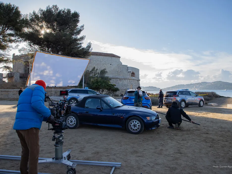 Cette photo montre un tournage en extérieur près de la plage de Pipady : une voiture est filmée par une équipe technique, avec caméra sur rails, réflecteur, et plusieurs véhicules de police en arrière-plan, devant la Tour royale. L’ensemble évoque une scène de film ou de série policière en préparation.