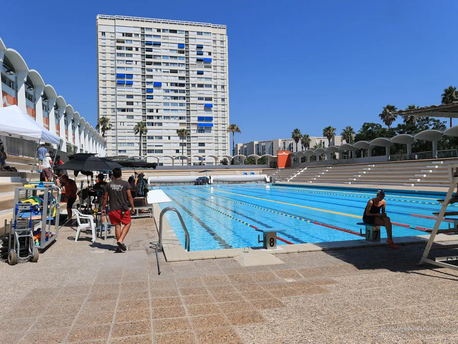 Cette image montre les préparatifs ou le déroulement d'un tournage d'un épisode de la série "Tom et Lola" dans le bassin extérieur du stade nautique du Port-Marchand, sous un ciel bleu dégagé.